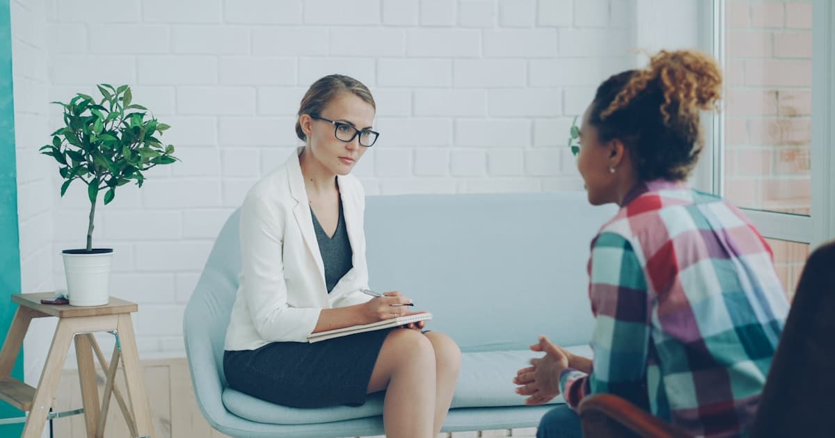 A therapist listens attentively to a client during a counseling session, taking notes on a clipboard in a bright, modern office
